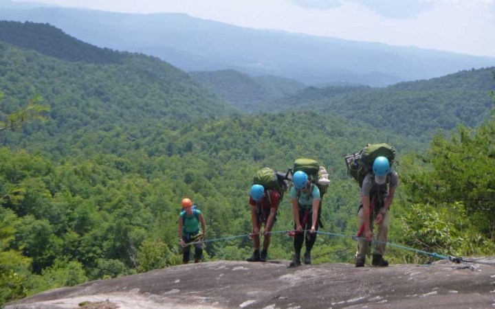 A group of students wearing helmets and backpacks hold on to a rope as they scramble up a rocky slope. Behind them is a green mountainous landscape. 